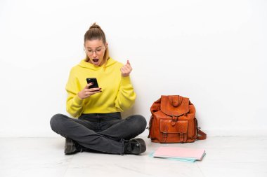 Beyaz arka planda beyaz bir zeminde oturan genç beyaz kadın şaşırdı ve bir mesaj gönderiyor.