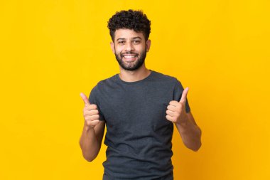 Faslı genç adam sarı arka planda izole edilmiş. Baş parmakları havada ve gülümsüyor.