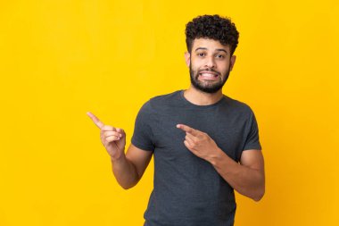 Faslı genç adam sarı arka planda izole edilmiş korkmuş ve kenarı işaret ediyor.