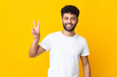 Sarı arka planda izole edilmiş genç Faslı adam gülümsüyor ve zafer işareti gösteriyor