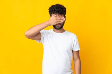 Faslı genç bir adam sarı arka planda izole edilmiş. Gözlerini elleriyle kapatıyor. Bir şey görmek istemiyorum.