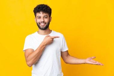 Faslı genç bir adam sarı arka planda izole edilmiş. Elinde bir ilan vermek için avucunda hayali bir alan tutuyor.