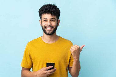 Genç Faslı adam mavi arka planda cep telefonu kullanarak ve yan tarafı işaret ederek izole edilmiş.