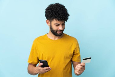 Genç Faslı bir adam mavi arka planda izole edilmiş cep telefonuyla kredi kartı satın alıyor.