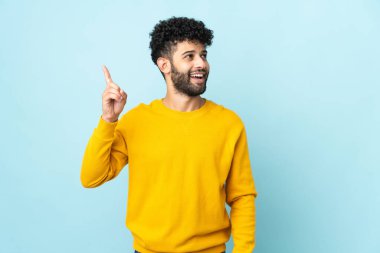 Faslı genç bir adam mavi arka planda izole edilmiş bir şekilde parmağını kaldırarak çözümü bulmaya çalışıyor.