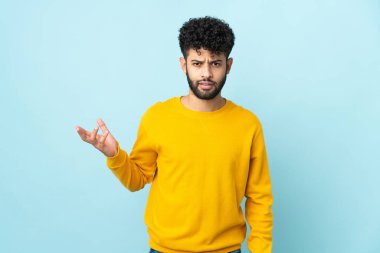 Faslı genç bir adam mavi arka planda izole edilmiş şüpheli hareketlerde bulunuyor.