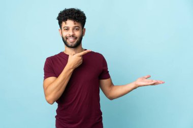 Faslı genç bir adam mavi arka planda izole edilmiş. Avucunda hayali bir şey tutuyor. Bir ilan vermek için.