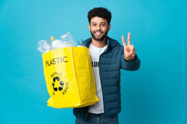 Genç Faslı bir adam, izole edilmiş arka planda geri dönüşüm için plastik şişelerle dolu bir çanta tutuyor. Gülümsüyor ve zafer işareti gösteriyor.