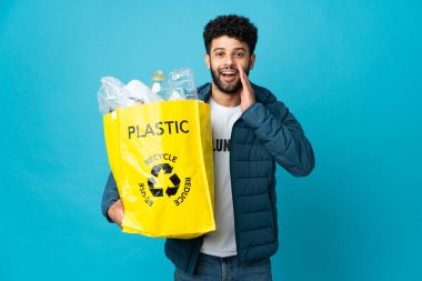 Bir çanta dolusu plastik şişeyi elinde tutan genç Faslı adam izole edilmiş arka planı şaşırmış ve şok olmuş bir yüz ifadesiyle geri dönüştürmek için.