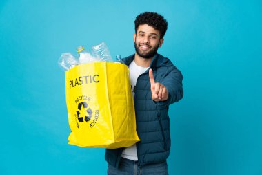Genç Faslı bir adam, izole edilmiş arka plan üzerinde geri dönüşüm yapmak ve parmağını kaldırmak için plastik şişelerle dolu bir çanta tutuyor.