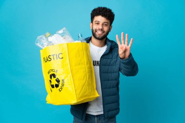 Genç Faslı bir adam izole edilmiş arka planda geri dönüşüm yapmak için plastik şişelerle dolu bir çanta tutuyor mutlu ve parmaklarıyla dört kişi sayıyor.