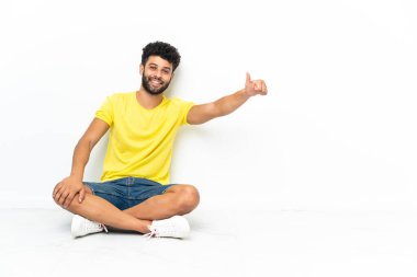 Genç Faslı yakışıklı adam, izole edilmiş arka planda yerde oturuyor ve baş parmak işareti yapıyor.