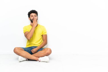 Genç Faslı yakışıklı adam izole bir zeminde oturuyor esniyor ve ağzı açık bir şekilde elleriyle ağzını kapatıyor.