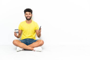 Genç Faslı yakışıklı adam izole edilmiş arka planda oturuyor. Elinde kahve ve cep telefonu var.