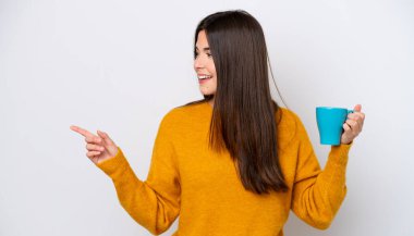 Genç Brezilyalı bir kadın beyaz arka planda izole edilmiş bir fincan kahve tutuyor ve bir ürünü sunmak için yan tarafa bakıyor.