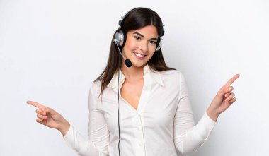 Beyaz arka planda izole edilmiş kulaklıkla çalışan beyaz pazarlamacı kadın parmak uçlarını işaret ediyor ve mutlu.