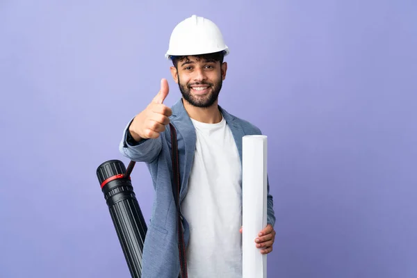 Young architect Moroccan man with helmet and holding blueprints over isolated background with thumbs up because something good has happened