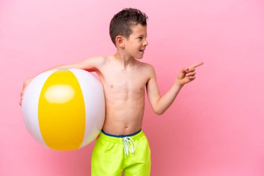 Pembe arka planda izole edilmiş bir plaj topu tutan küçük beyaz çocuk bir ürünü sunmak için yan tarafı işaret ediyor.