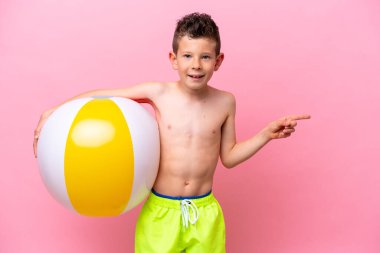 Pembe arka planda izole edilmiş bir plaj topu tutan küçük beyaz çocuk şaşırdı ve parmağını yana çevirdi.