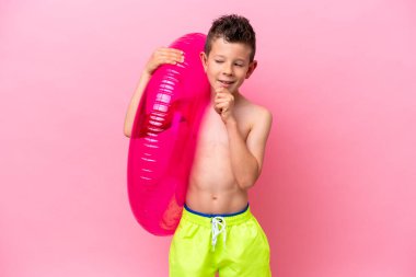 Küçük beyaz çocuk pembe arka planda izole edilmiş şişirilebilir bir çörek tutuyor. Yan tarafa bakıyor ve gülümsüyor.