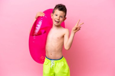 Küçük beyaz çocuk pembe arka planda izole edilmiş şişirilebilir bir çörek tutuyor gülümsüyor ve zafer işareti gösteriyor.