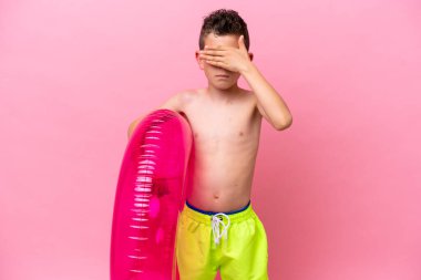 Küçük beyaz bir çocuk pembe arka planda izole edilmiş şişirilebilir bir çörek tutuyor. Gözlerini elleriyle kapatıyor. Bir şey görmek istemiyorum.