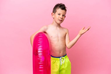 Küçük beyaz çocuk pembe arka planda izole edilmiş şişirilebilir bir çörek tutuyor. Davet etmek için ellerini yana uzatıyor.