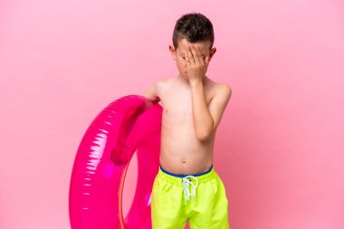 Küçük beyaz çocuk elinde şişme bir çörek tutuyor. Pembe arka planda, yorgun ve hasta bir ifadeyle izole edilmiş.