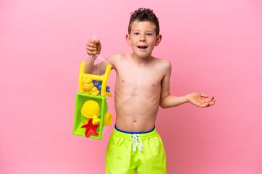 Küçük beyaz çocuk plaj oyuncakları tutuyor. Pembe arka planda izole edilmiş. Yüzünde şok ifadesi var.