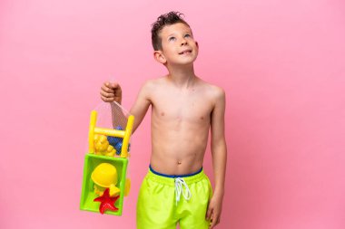 Küçük beyaz çocuk pembe arka planda izole edilmiş plaj oyuncakları tutuyor gülümserken yukarı bakıyor.