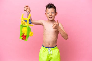 Küçük beyaz çocuk pembe arka planda izole edilmiş plaj oyuncakları tutuyor. Başparmakları havada çünkü iyi bir şey oldu.