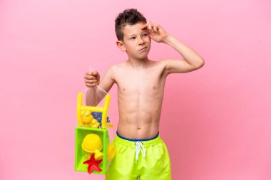 Pembe arka planda izole edilmiş plaj oyuncakları tutan küçük beyaz çocuk şüpheleri ve kafa karıştırıcı bir ifadeyle