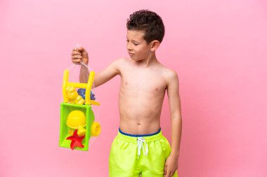 Küçük beyaz çocuk elinde plaj oyuncakları tutarken üzgün bir ifadeyle pembe arka planda izole edilmiş.