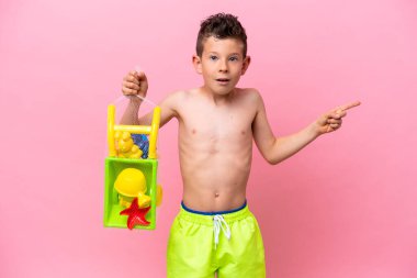 Pembe arka planda izole edilmiş plaj oyuncakları tutan küçük beyaz çocuk şaşırmış ve parmağıyla kenarı işaret ediyor.