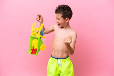 Pembe arka planda plaj oyuncakları tutan küçük beyaz çocuk zaferi kutluyor.