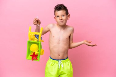 Küçük beyaz çocuk pembe arka planda plaj oyuncakları tutuyor. Omuzlarını kaldırırken şüphe uyandırıyor.