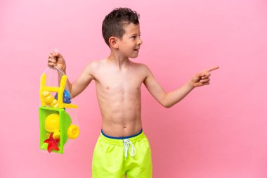 Pembe arka planda izole edilmiş plaj oyuncakları taşıyan küçük beyaz çocuk bir ürünü sunmak için kenarı işaret ediyor.