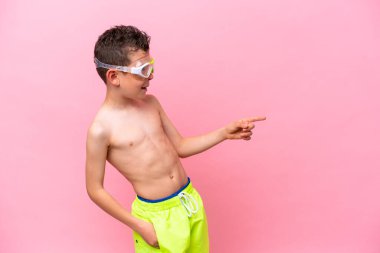Pembe arka planda izole edilmiş dalış gözlüğü takan beyaz bir çocuk parmak göstererek bir ürün sunuyor.