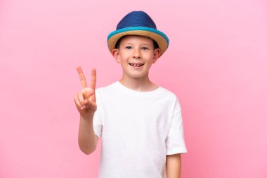 Pembe arka planda izole edilmiş şapka takan küçük beyaz çocuk gülümsüyor ve zafer işareti gösteriyor.