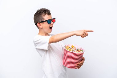 Little caucasian boy isolated on white background with 3d glasses and holding a big bucket of popcorns while pointing away
