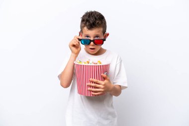 Beyaz arka planda izole edilmiş küçük beyaz çocuk 3D gözlüklü ve elinde koca bir kova patlamış mısırla şaşırdı.