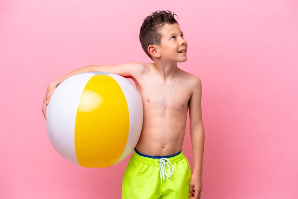 一个拿着一个粉色背景的海滩球的高加索小男孩微笑着抬起头来 — 图库照片