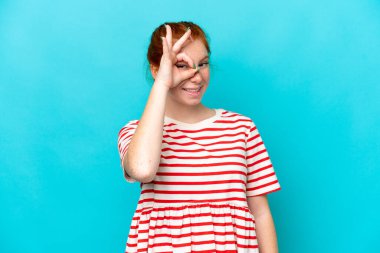 Mavi arka planda izole edilmiş genç kızılımsı kadın parmaklarıyla tamam işareti gösteriyor.