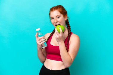 Kırmızımsı genç bir kadın mavi arka planda bir elma ve bir şişe suyla izole edilmiş.