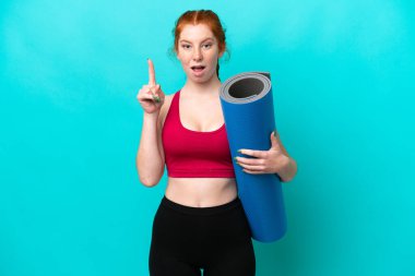 Genç spor kırmızısı kadın yoga derslerine giderken elinde mavi arka planda izole edilmiş bir minder tutarken parmağıyla işaret eden bir fikir düşünüyor.