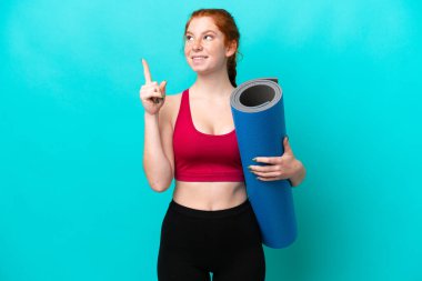 Genç spor kırmızısı kadın yoga derslerine giderken elinde mavi arka planda izole edilmiş bir minder tutarak harika bir fikre işaret ediyor.