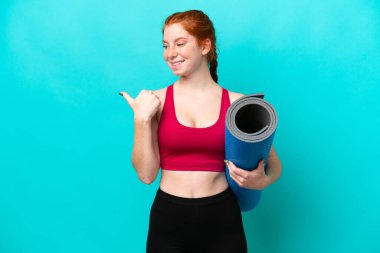 Genç spor kırmızısı kadın yoga derslerine giderken elinde mavi arka planda izole edilmiş bir minder tutarak bir ürünü sunmak için yan tarafı işaret ediyor.