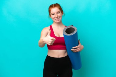 Genç spor kırmızısı kadın yoga derslerine giderken mavi arka planda bir minder tutuyor. Başparmakları havada, çünkü iyi bir şey oldu.