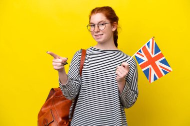Young hispanic woman holding an United Kingdom flag isolated on yellow background pointing finger to the side
