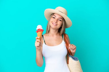 Elinde dondurma ve plaj çantası olan genç Romen kadın mavi arka planda izole edilmiş gülümsüyor.